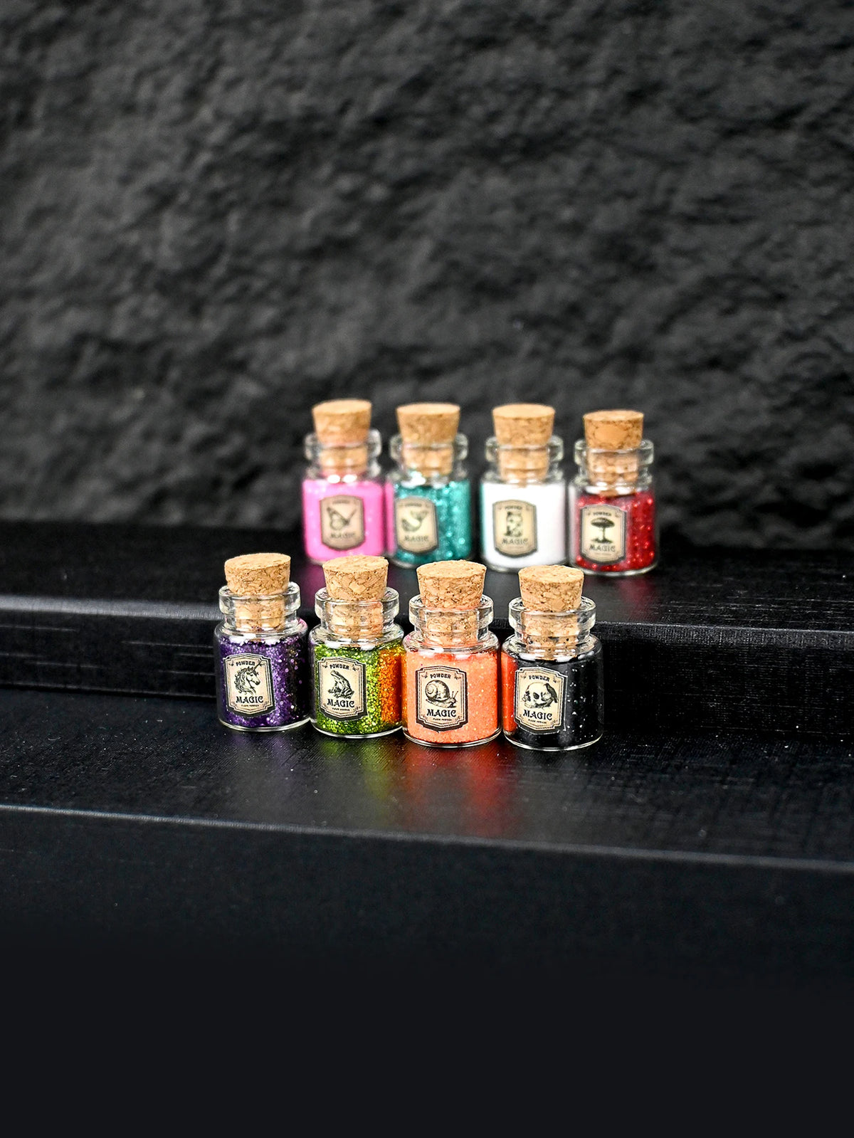 A collection of small glass bottles filled with colorful glitter powder for party decorations arranged on a dark surface.