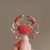 A close-up of a vintage marine life brooch pin set featuring a vibrant red crab design being held by a hand.