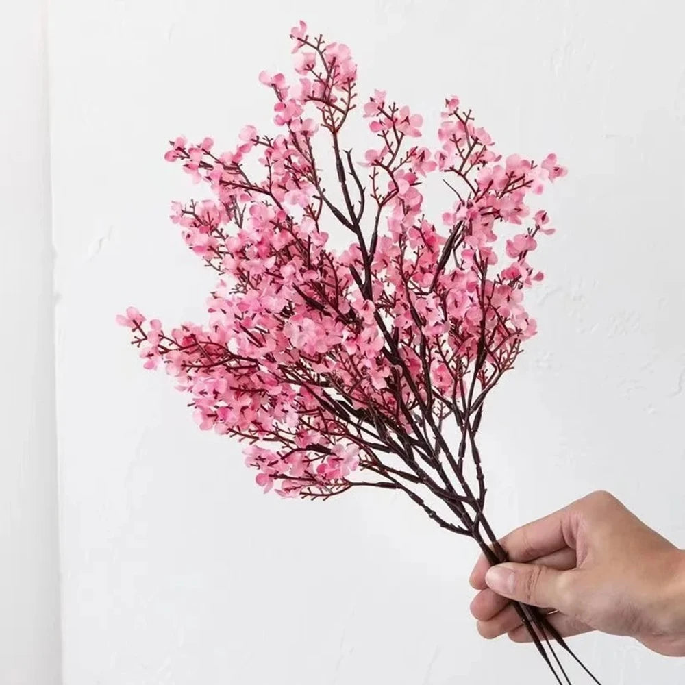 A hand holds a branch of pink cherry blossom and gypsophila artificial flowers against a white background.