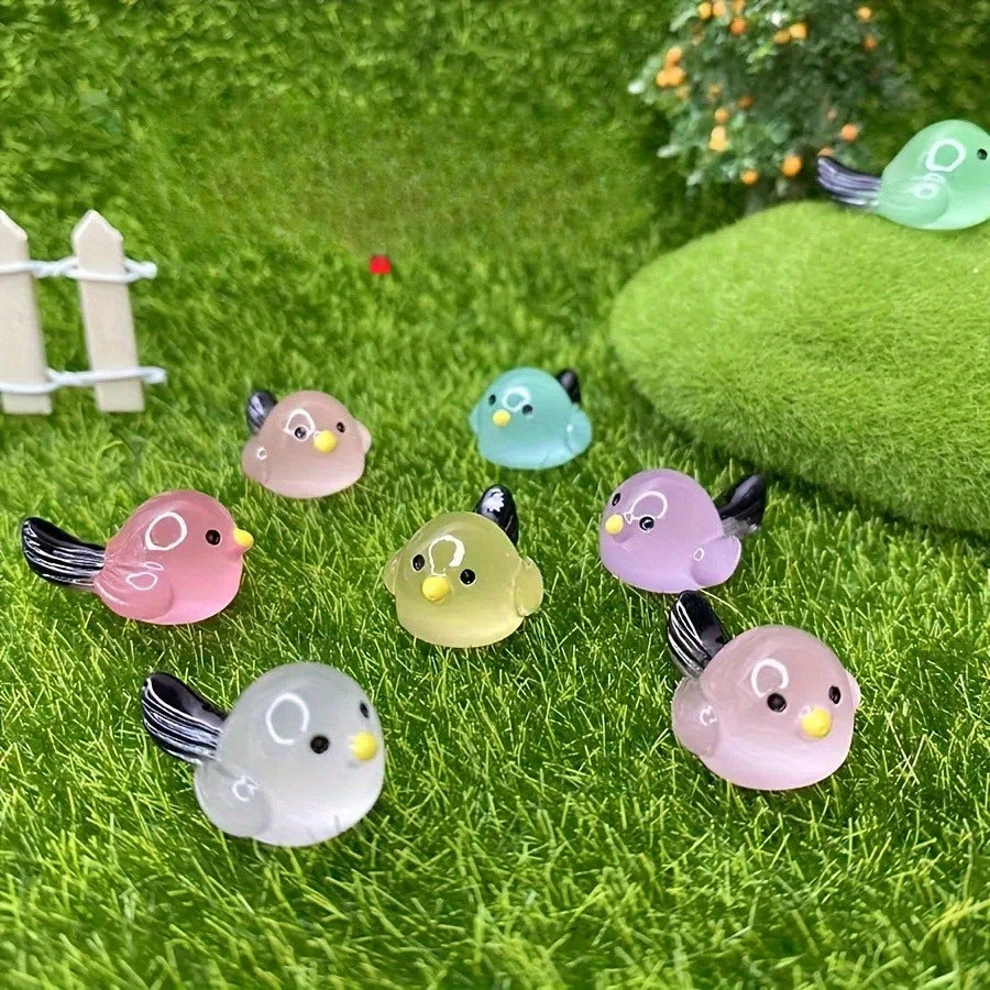 A set of small luminous resin bird fairies in various pastel colors placed on green grass with moss and foliage.