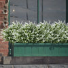 A dense planting of white artificial flowers in a green planter box set against a brick wall and glass window.