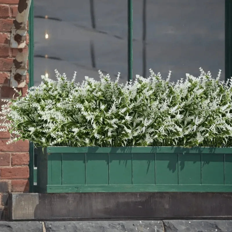 A dense planting of white artificial flowers in a green planter box set against a brick wall and glass window.