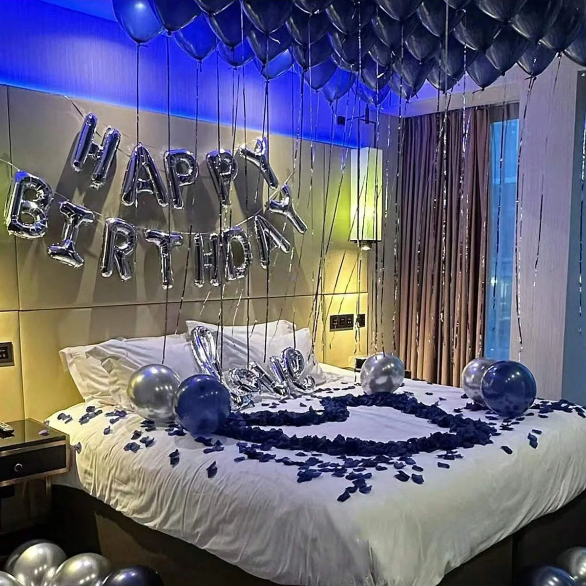 Bett dekoriert mit dunkelblauen Ballons, die 'Happy Birthday' bilden, und verstreute blaue Blütenblätter.
