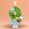 A green glitter number eight candle decorated with a white daisy and a bee, set on a white base.