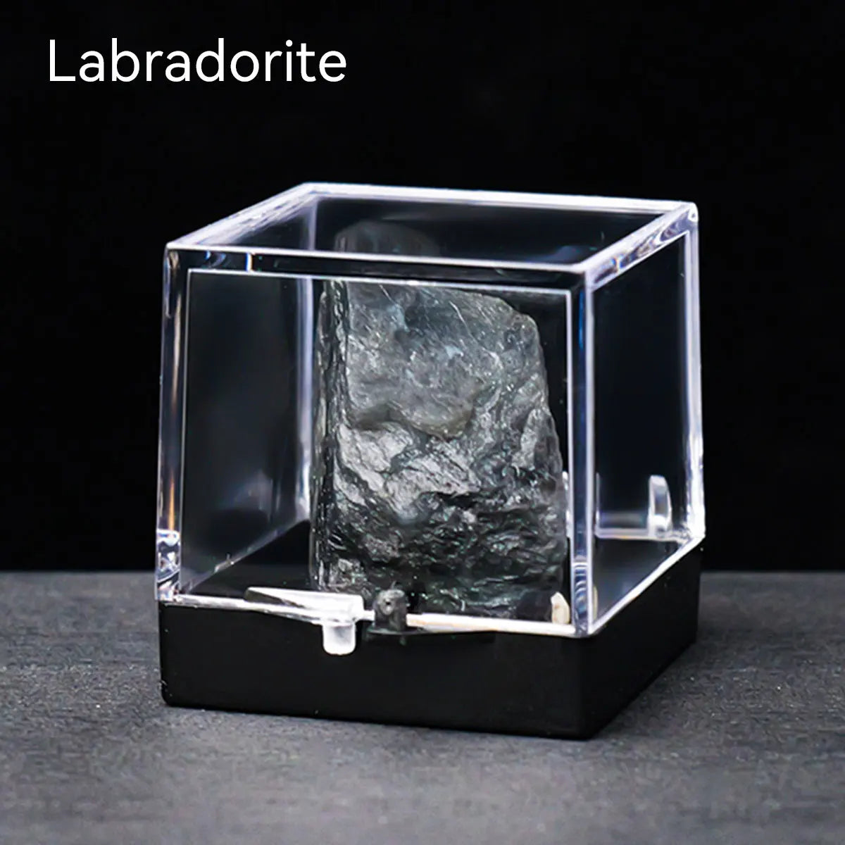 A clear glass box displays a rough, dark gray labradorite crystal specimen on a black surface.