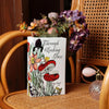 A white ceramic book-shaped vase filled with dried flowers sits on a woven surface next to a wooden chair.