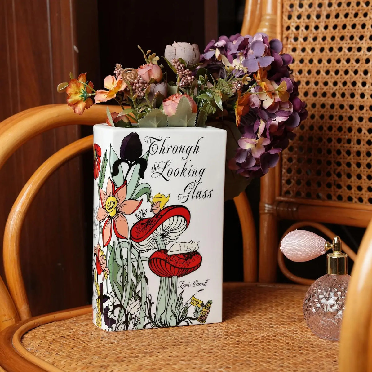 A white ceramic book-shaped vase filled with dried flowers sits on a woven surface next to a wooden chair.