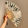 A hand is holding an open folding fan with a leopard print design and black handle.