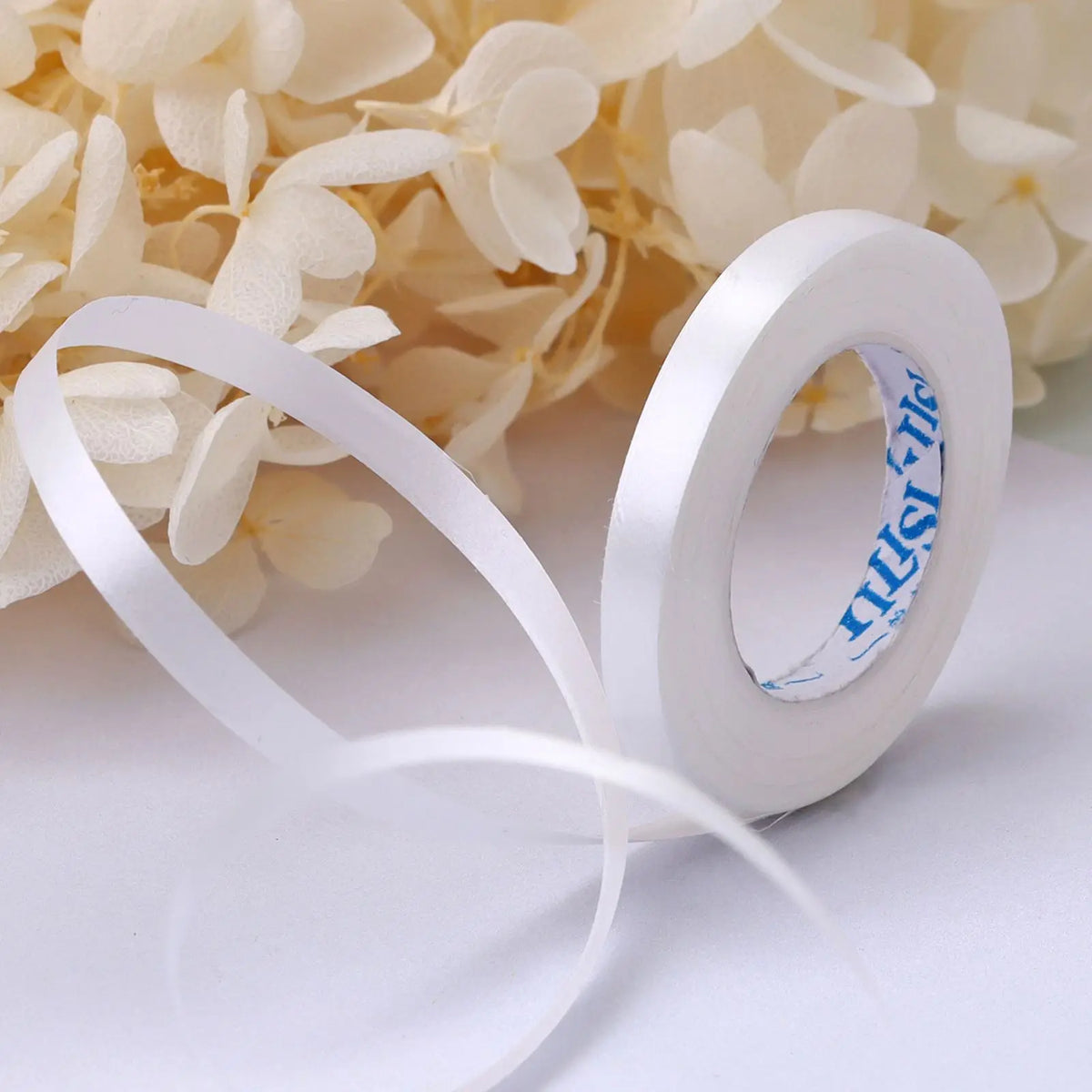 A roll of white satin balloon ribbon with blue text is shown next to a cluster of light yellow flowers.
