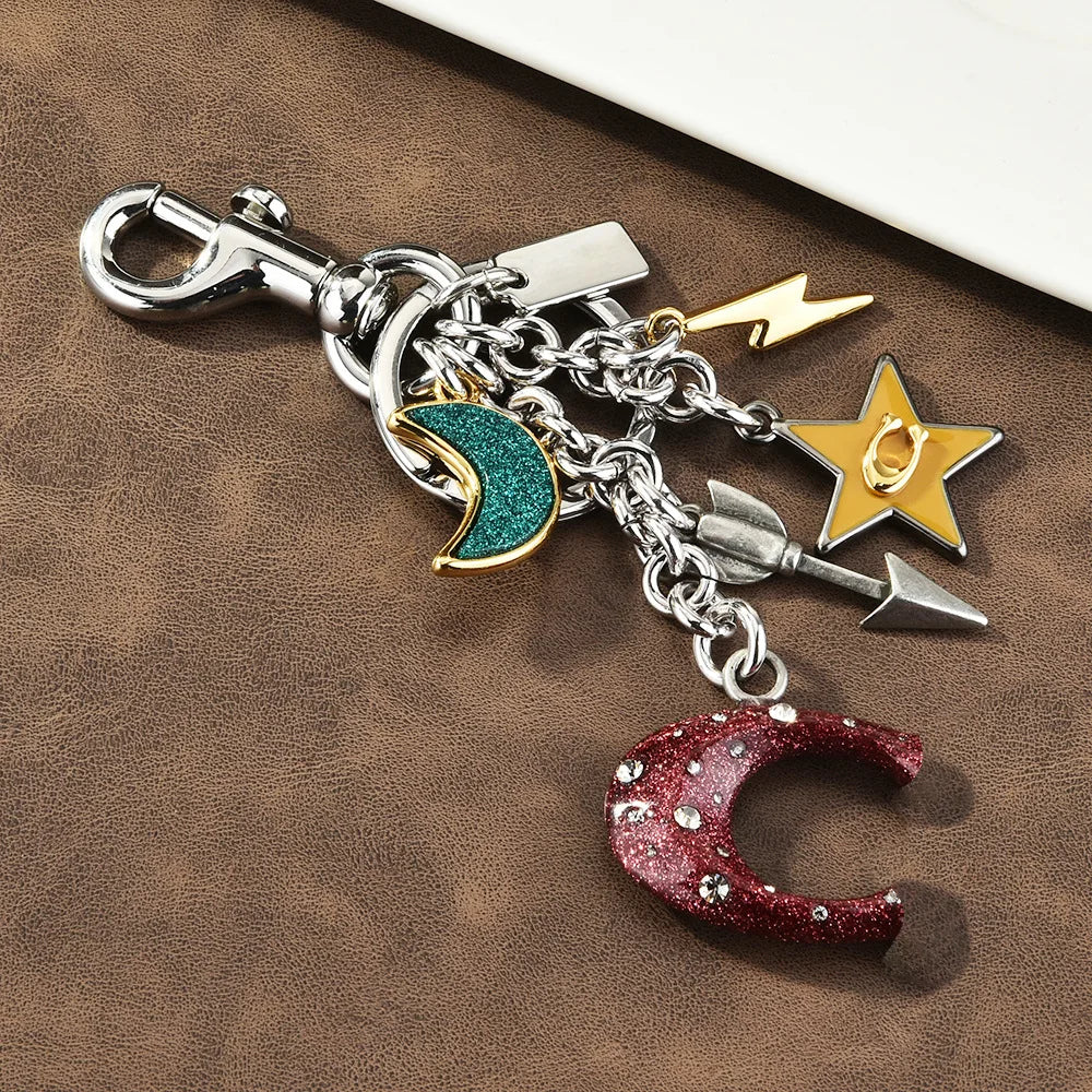 Silver keychain featuring a crescent moon charm, a gold star charm, and an arrow charm on a leather background.