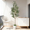 A tall artificial olive tree branches in a white pot displayed next to a white cabinet in a bright room.