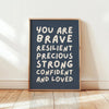 A framed canvas print with white text on a dark blue background that reads You are brave resilient precious strong confident 