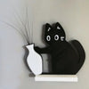 Black acrylic cat figurine with long whiskers and a white vase on a light gray wall.
