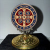 A round metal ornament featuring Latin symbols and religious iconography on a gold base.