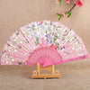 Pink silk flower folding fan with white and purple floral patterns displayed on a wooden stand.