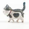 A small, white and gray cartoon cat figurine with a dark tail and a brown collar.