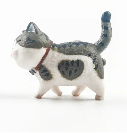 A small, white and gray cartoon cat figurine with a dark tail and a brown collar.