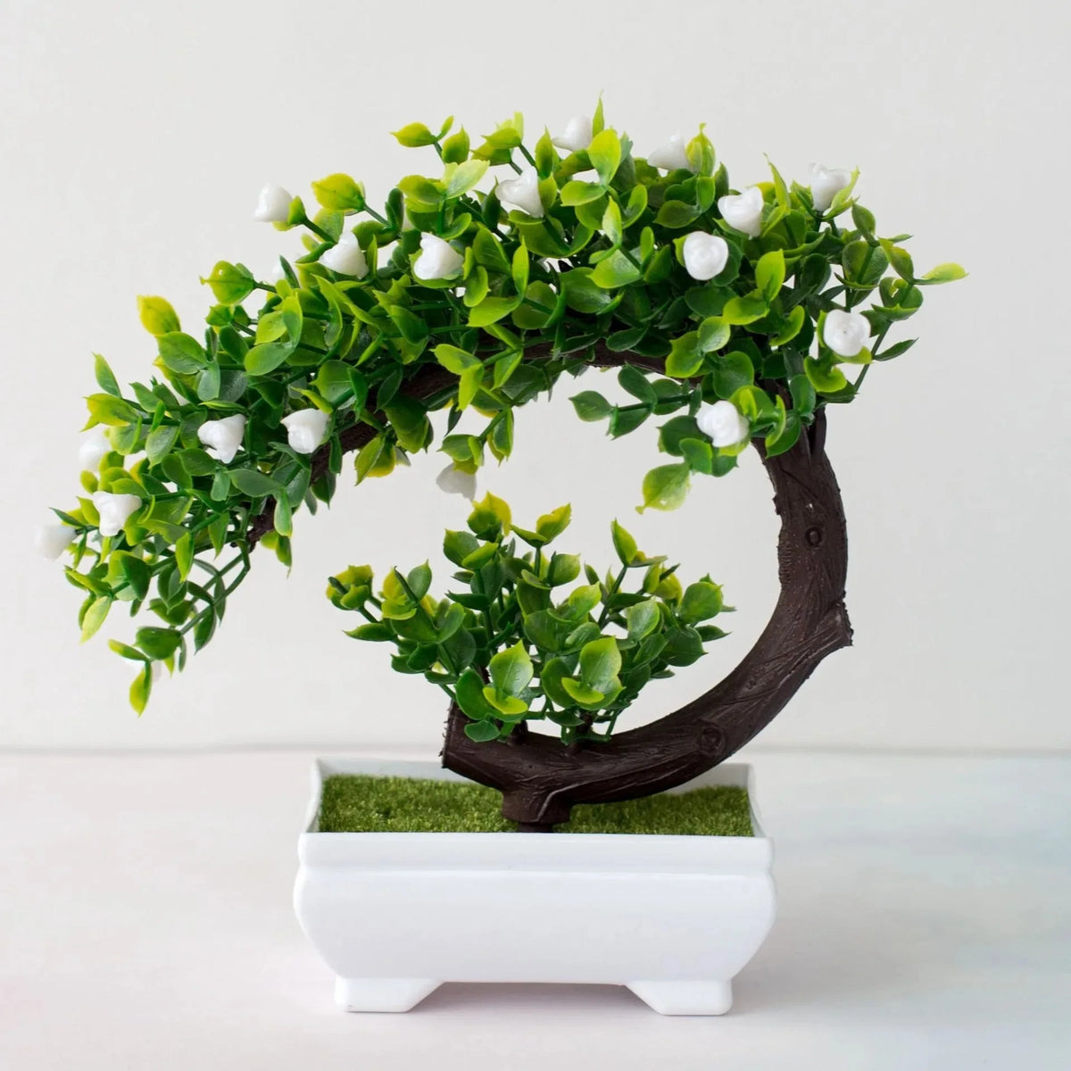 An artificial bonsai tree decoration with bright green leaves and white blossoms growing from a dark brown trunk in a white p