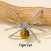 Silver crystal spider figurine with a golden tiger eye bead resting on a natural wood surface.