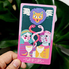 A tarot card featuring a lion head and two figures in soft pink and sky-blue artwork.