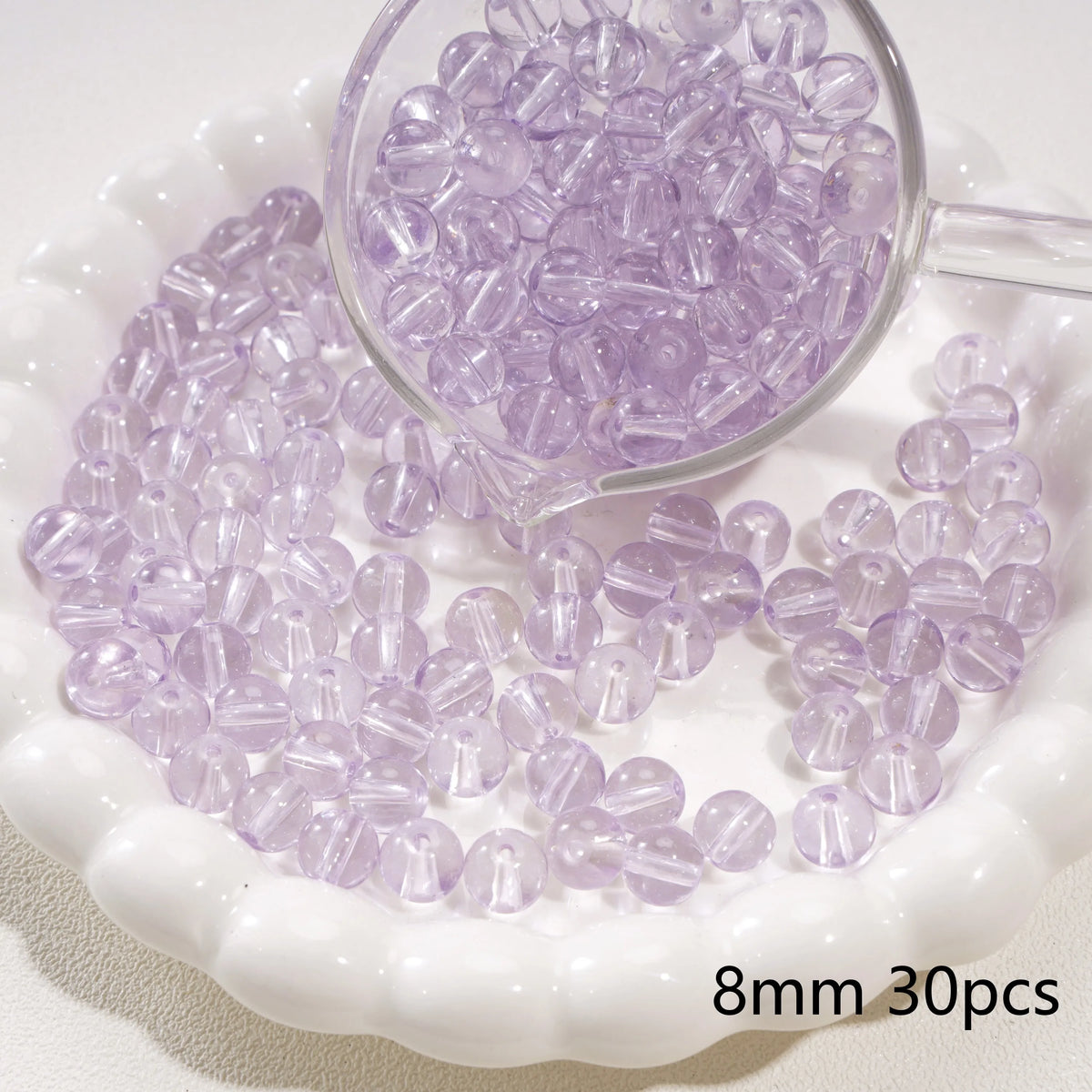 A clear glass measuring spoon holds purple spacer beads scattered on a white dish.