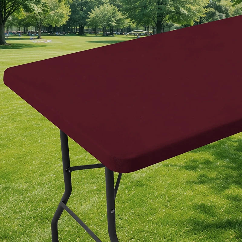 A maroon stretch tablecloth is displayed on a folding table, set in a grassy outdoor area.
