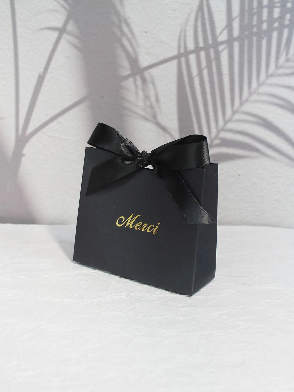 A black paperboard thank you gift bag tied with a black ribbon and gold script reading Merci.