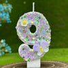 A purple glitter number cake candle shaped like the number 8 decorated with a white flower and a purple butterfly