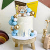 A white cake decorated with blue and gold accents, featuring a bear and star topper on a wooden stand.
