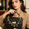 A woman holds a black quilted handbag with gold chain charms against a beige blazer.