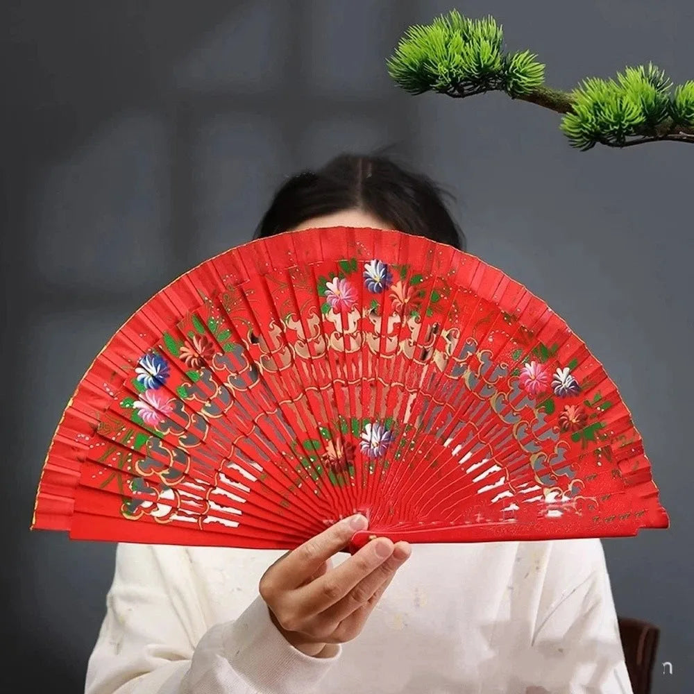 A hand-painted red folding fan with floral designs is held by a person against a gray background.