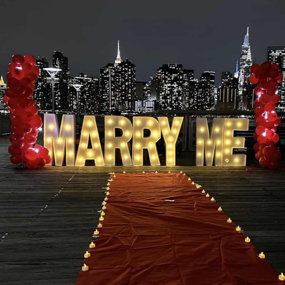 A marquee light-up sign spelling out 'MARRY ME' decorated with red balloons and a red carpet leading toward a city skyline at