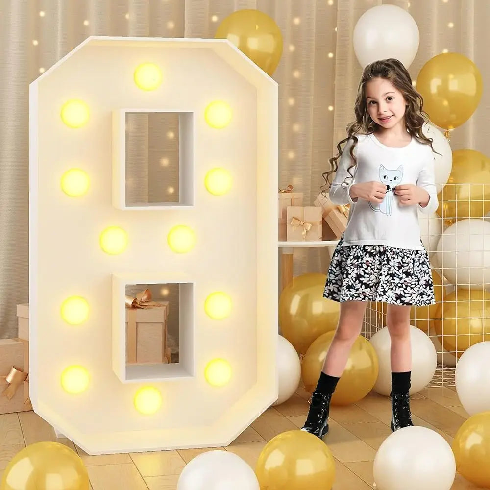 A young girl stands next to a large white LED light-up number decoration shaped like a house, surrounded by yellow and white 