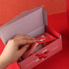 A hand is opening a pink gift box with a lid, revealing a red interior and a surprise message.