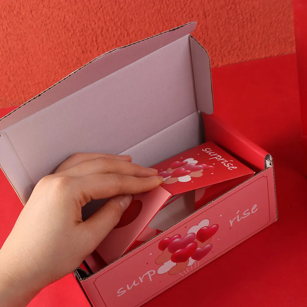 A hand is opening a pink gift box with a lid, revealing a red interior and a surprise message.