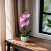 A realistic artificial orchid potted plant with pink flowers sits on a wooden table next to a window.