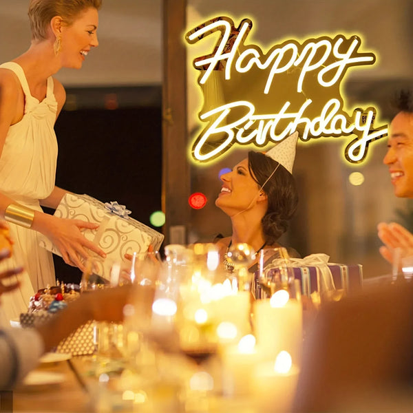 A woman smiling while a neon sign reads Happy Birthday in yellow light on a wooden table.