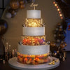 A four-tiered white cake display featuring orange and pink roses illuminated by warm LED lights.