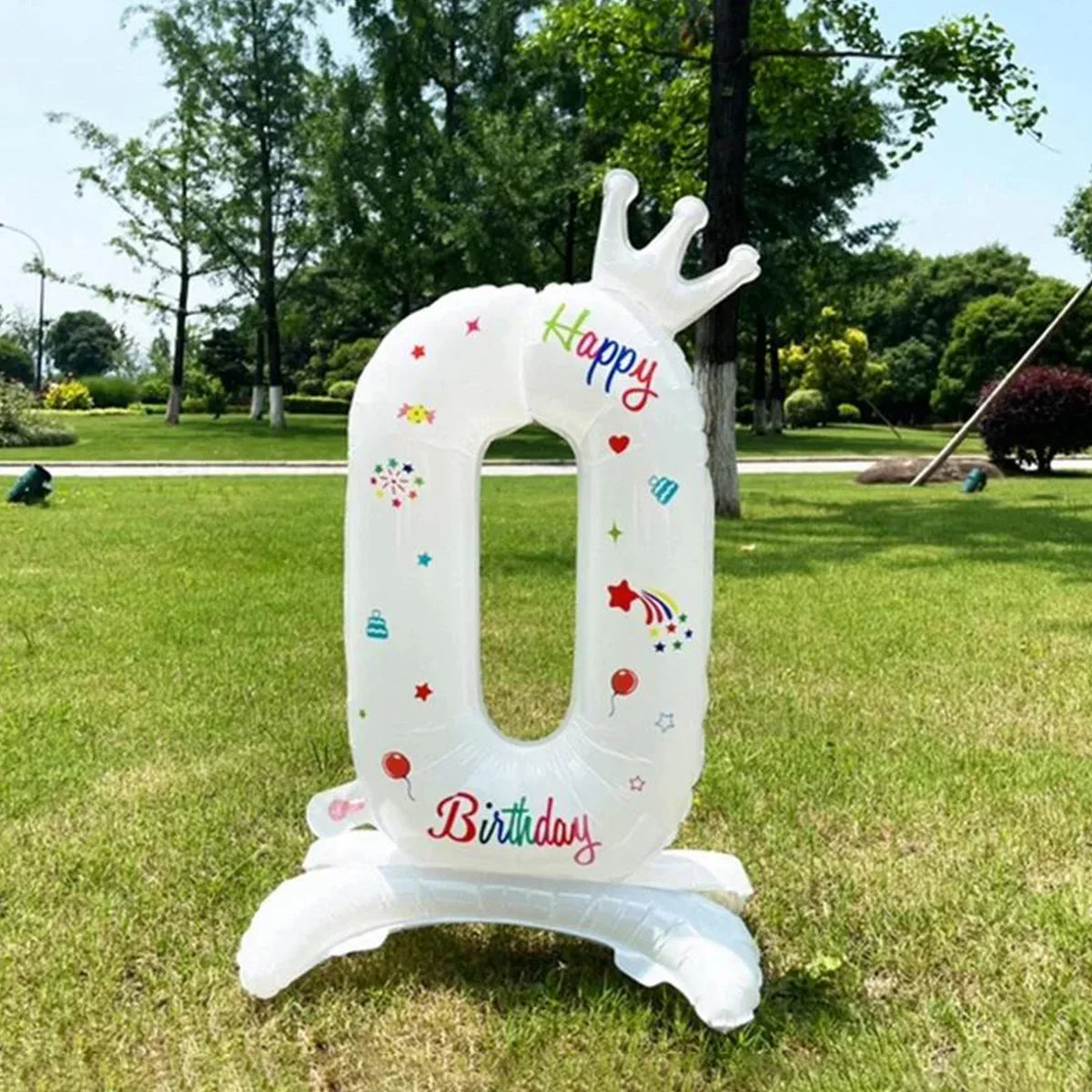 A white foil balloon shaped like the number 0 with a crown and birthday decorations standing on the grass.