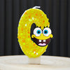 A yellow sponge cake candle shaped like the number 0, decorated with orange glitter and a SpongeBob face.