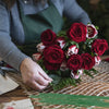 A hand places deep red roses in a green floral wire arrangement on a wooden surface.