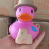 A pink rubber duck bath toy with an orange beak and a gray hat is held in a hand.