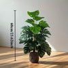 A lush green artificial monstera palm plant in a pot displayed against a white wall with a measuring stick.