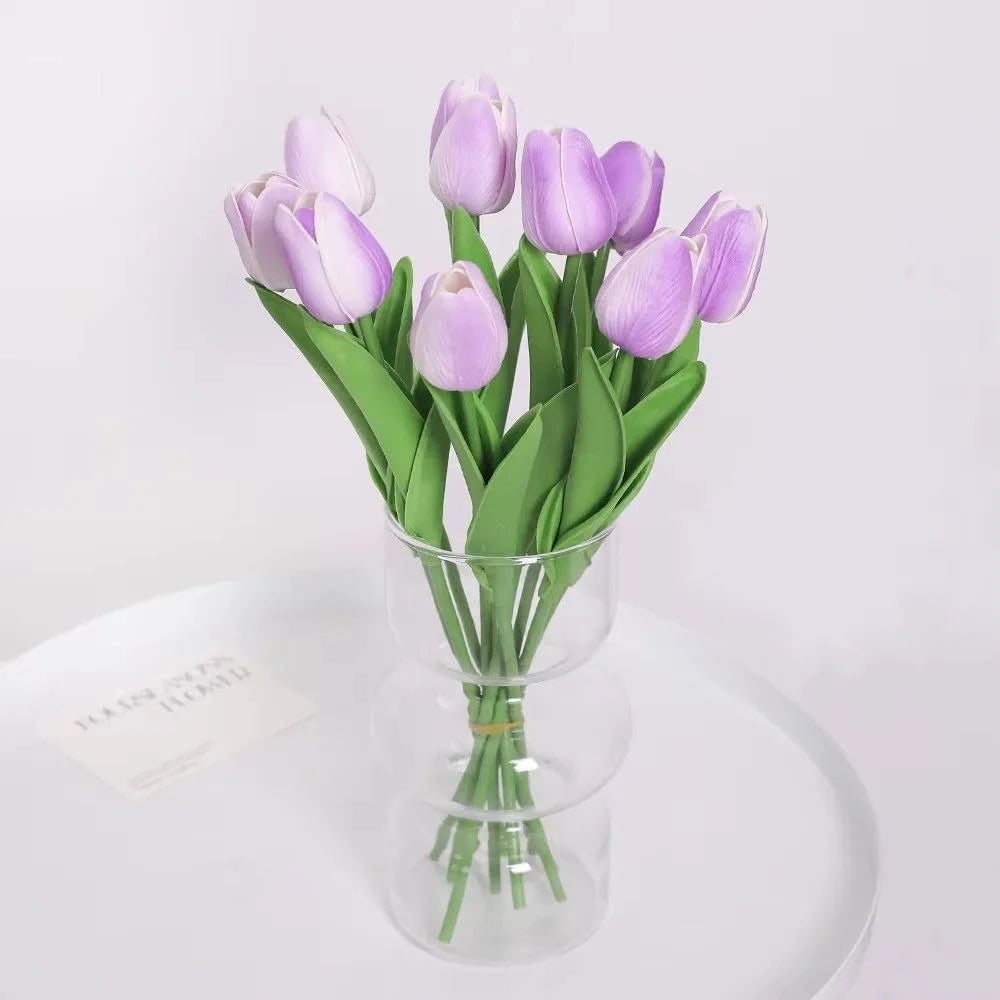 A bouquet of artificial purple tulips in a clear glass vase on a white surface.