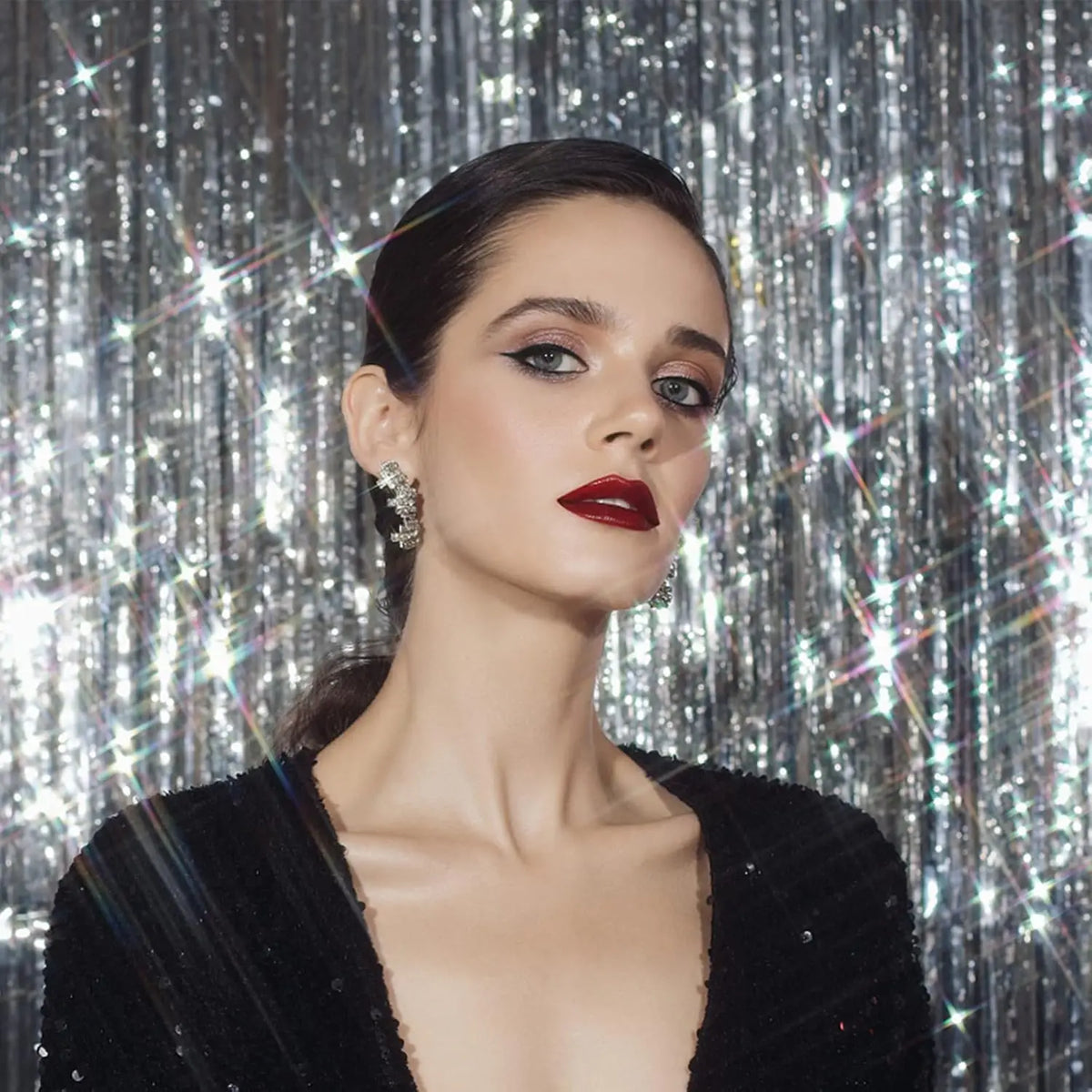 A woman with dark hair and dramatic makeup looks toward the camera against a silver metallic foil fringe curtain backdrop.