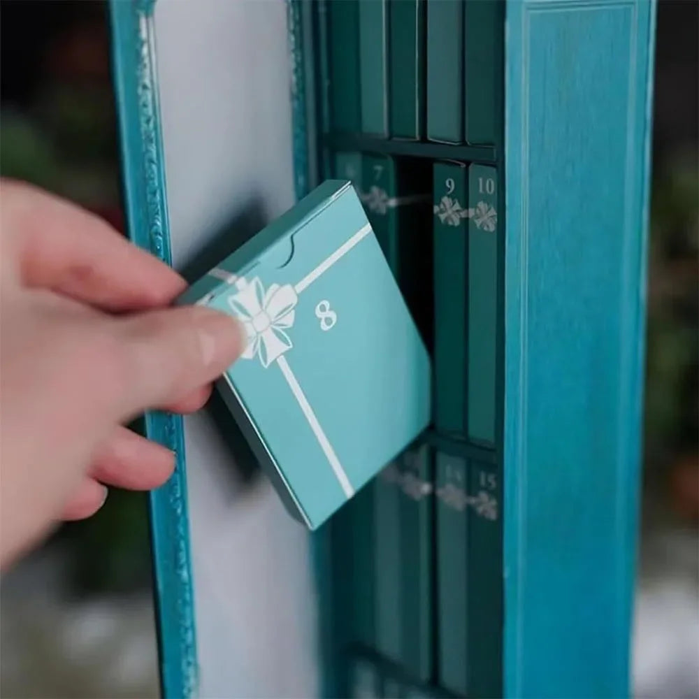 A hand is opening a teal-colored advent calendar box with a white ribbon and ornaments inside.
