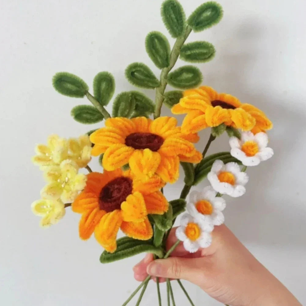 A hand holds a bouquet of multi-colored chenille stem pipe cleaners featuring yellow, orange, and white flowers.