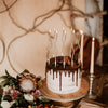 A dark chocolate birthday cake decorated with dripping ganache and lit candles on a wooden stand.