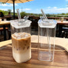 A clear glass coffee mug filled with iced latte sits on a wooden table outdoors.