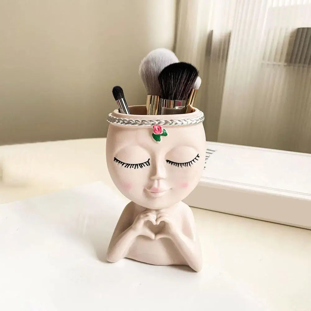 A small artistic resin girl face planter with painted features and a brush holder on a white surface.